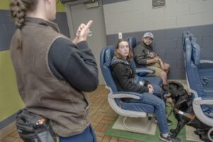 A woman stands in front of two clients who are sitting in airplane chairs with their dogs at their feet at the downtown Rochester training facility. 