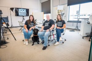 Group photo of three people and a black Labrador in a modern office setting, featuring a Taking the Lead logo on a screen. The room has studio lights and a whiteboard, suggesting a professional environment.