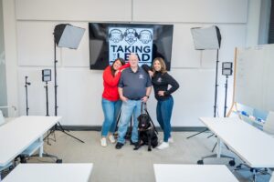 Group of three people and a guide dog posing in a modern conference room. They stand in front of a screen displaying a logo with the text Taking the Lead. Two women wear casual attire, smiling alongside a man holding the dog's harness. Professional lighting equipment and a whiteboard are visible in the background.