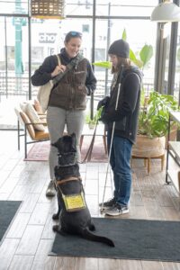 Two women stand in a bright lobby full of windows. Two chairs and a few plants are in the background.