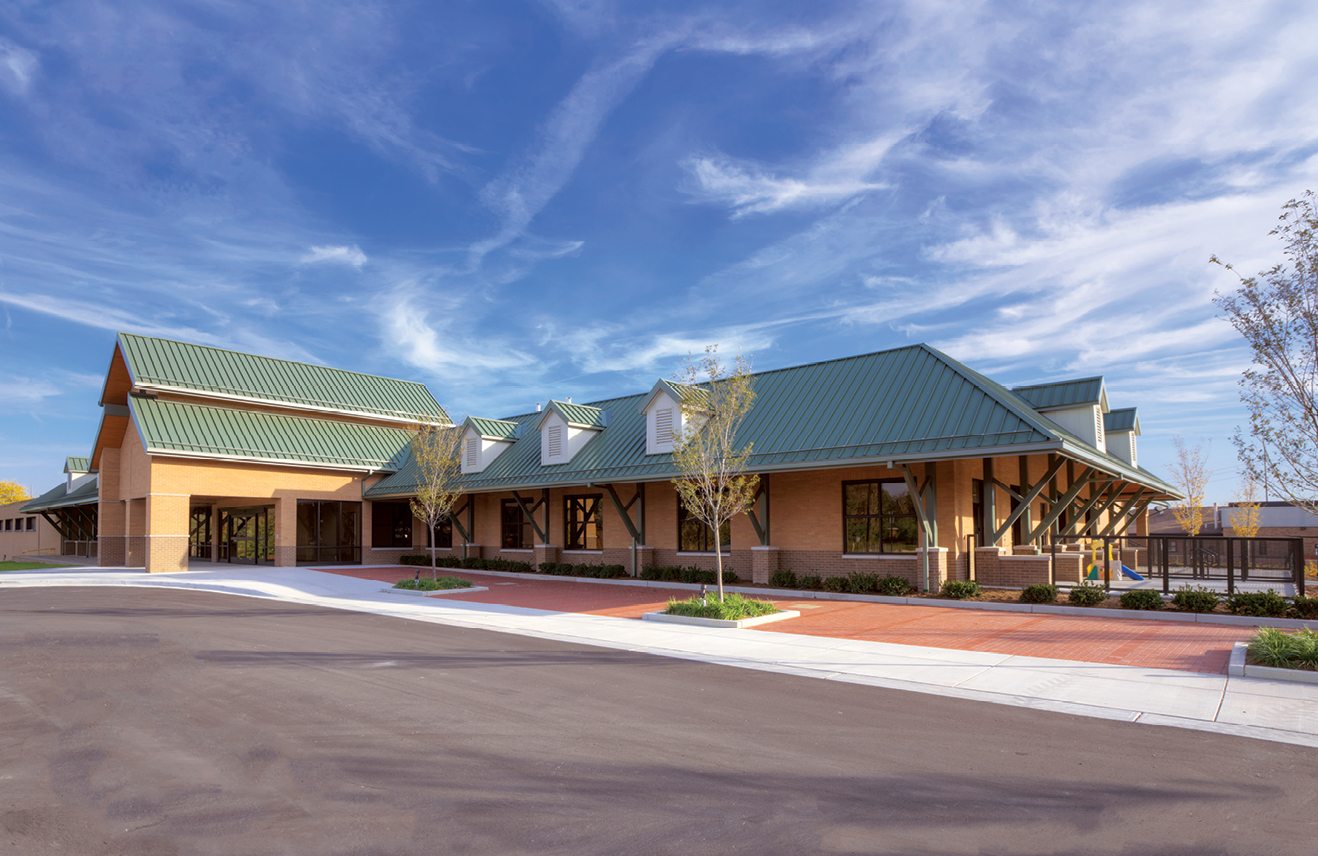 A modern building with a green metal roof and brick facade is set against a clear blue sky, featuring landscaped pathways and trees.