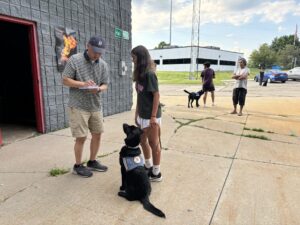 Man and teenage girl with a Leader Dog in training outside a building while another person walks by with a service dog in the background – sunny day, training session.