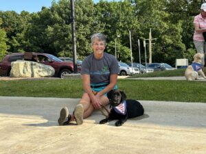 A woman in a grey t-shirt and shorts smiles while sitting on concrete beside a black puppy wearing a Future Leader Dog bandana during a training event at a park. Lush green trees and parked cars form the background with another puppy in training and handler visible in the distance.