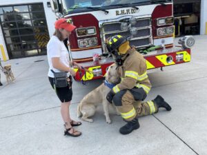 A firefighter in full gear kneels down next to a golden retriever wearing a blue Future Leader Dog bandana. The dog is smiling up at the firefighter. A woman stands next to the dog looking at the exchange while holding onto the dog's leash.
