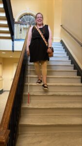 A woman uses her white cane to navigate a stairwell inside a stone building. She is wearing a black and pink outfit and is smiling widely.
