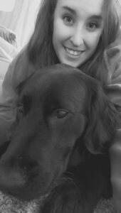 A woman with long wavy hair smiles at the camera. In front of the person is a black Labrador and golden retriever cross. The image is in black and white.