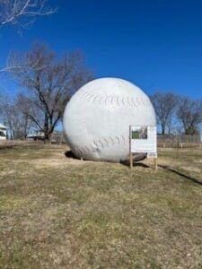 A large, round structure designed to look like a baseball, with stitches details, placed in a grassy field under a clear blue sky.