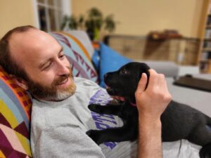 A man sits on a couch holding a black Labrador retriever puppy on his chest smiling.