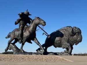 A large, bronze statue of Buffalo Bill Cody depicts Cody holding a rifle while riding his running horse and is riding beside and pointing the rifle at a large, running buffalo to his left.