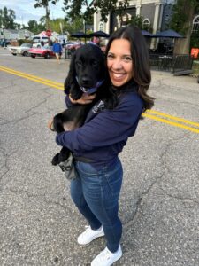 A person with dark mid-length hair is standing in a street during a vintage car event. She is smiling toward the camera while holding a young black Labrador retriever.