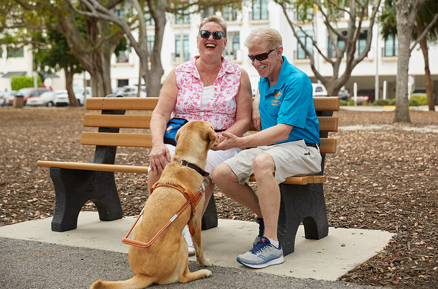 A couple sits on a bench in a park, laughing and interacting with a dog at their feet.