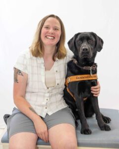 Smiling woman sitting with a black Labrador wearing a harness labeled "LeaderDog.org".