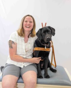 Smiling woman sitting with a black Labrador wearing a harness labeled "LeaderDog.org". The woman is leaning into the dog smiling widely while holding up bunny fingers behind the dogs head.