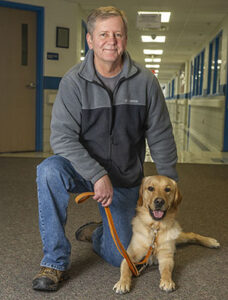 A man in zip-up fleece jacket and jeans kneels on a gray carpet next to a yellow lab lying next to him.