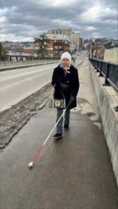 Elderly woman walking with a white cane on a city sidewalk in winter, wearing a black coat and white hat, with urban buildings and cloudy skies in the background.