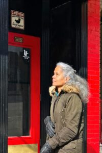 Person standing in front of a bright red building with a black door marked with a "VOTE HERE" sign. The individual is wearing a parka and looking to the side, bathed in sunlight.