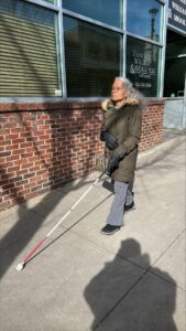 A woman walks along a sidewalk using a white cane. She is dressed in a heavy winter coat.