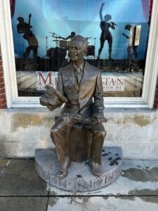 Bronze statue of a man sitting in front of a window with music-themed silhouettes, located in a downtown area.