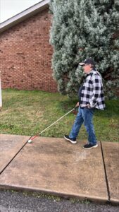 A person using a white cane walks along a sidewalk next to a brick building and a large evergreen bush.