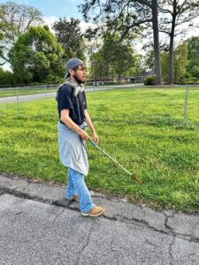 A young, bearded man wearing a backwards baseball cap, t-shirt and jeans, uses his cane to follow a grass line at the edge of the street.