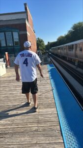 Man with a baseball jersey with the name "Tio Luis" written on it and wearing a ballcap walking on a train platform, holding a cane, with train approaching, sunny day.