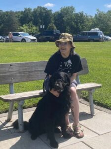 A woman wearing a bucket hat sits on a park bench on a sunny day. Sitting in front of her is a black Lab/golden retriever cross dog.
