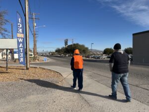Two people walking on a sidewalk in an urban area on a sunny day. One wears an orange reflective vest and the other a black beanie. A vertical sign beside them reads ICE AND WATER. The street is lined with trees and businesses in the background. The person in the orange vest is using a white cane.