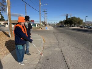Two people stand on the sidewalk at a crosswalk, with one person using a white cane. They are dressed warmly in winter clothing, including hats and an orange safety vest. The street is quiet with minimal traffic and clear skies. The setting is an urban environment with visible street signs and electrical poles.
