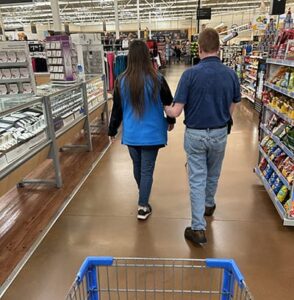 Man walking with woman in department store. The man is holding the woman's elbow as she acts as a sighted guide