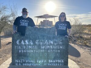 Two individuals stand in front of the Casa Grande National Monument sign, wearing matching Taking the Lead t-shirts. The prehistoric ruins and desert landscape are visible in the background under a bright, sunny sky.