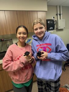 Two people smiling in a vet's office, each holding a small black puppy. They wear colorful hoodies with university logos.