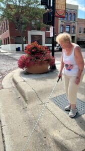 A woman uses her white cane to locate the curb at a downtown corner. She is wearing a white top and light pants.