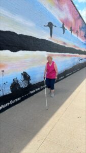 A woman uses her white cane on a wide sidewalk next to mural of sandhill cranes flying over water. She is wearing a bright top with dark pants.