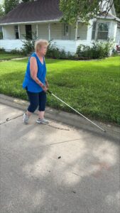 A woman uses her white cane to follow the curb while walking in the street. She is wearing a blue top and dark pants.