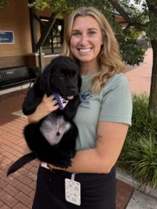 A young woman stands outside holding a black Labrador retriever puppy. The puppy is wearing a "Future Leder Dog" bandana.
