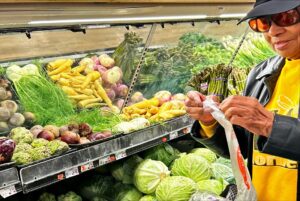 A person stands in the produce section of the grocery store, attempting to open a produce bag. They are wearing a dark hat, yellow shirt, and a dark coat.