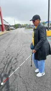 A person uses a support cane and long white cane in unison while crossing a parking lot. They wear sunglasses, a dark coat and jeans.
