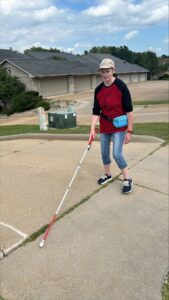 A woman using a white cane walks along a sidewalk near a driveway. 