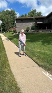A woman walks with a white cane on a sidewalk on a sunny day in a residential subdivision.