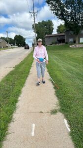 A woman walks along a sidewalk. She is using a white cane. Her cane is on the grass along the edge of the sidewalk, showing her the path.
