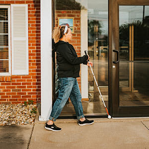 Woman in black hoodie and jeans walks with long white cane on a sidewalk in front of a brick building