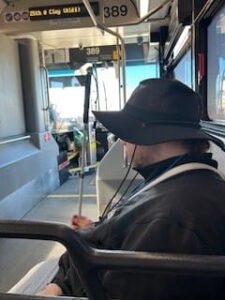 A person wearing a wide-brimmed hat and holds their white cane vertically while sitting on a bus.
