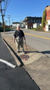 A person navigating a suburban street using two white canes. The person stands near a curb and a manhole cover on a sunny day, highlighting urban accessibility and mobility for those with visual impairments.