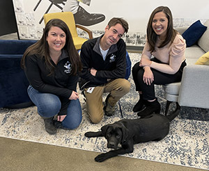 Christina and Leslie kneel next to Eric, and there's a black lab lying on the floor next to them