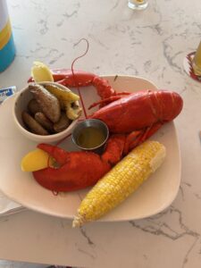 a bright red lobster sits on a white plate on a white table. It is accompanied by corn on the cob and a bowl of potatoes. The lobster has a lemon wedge between each claw.
