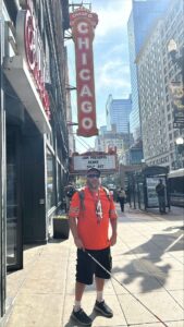 A man holding a white cane stands outside the iconic Chicago Theatre, with its bright red marquee sign, located in the bustling downtown area. The marquee announces a sold-out Keane concert presented by Jam. The street view includes urban architecture and people, typical of a lively Chicago scene.
