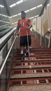 A person wearing an orange jersey and sunglasses uses a white cane to navigate down a set of stairs in a brightly lit indoor space, highlighting mobility and accessibility for visually impaired individuals.
