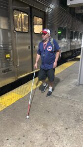 A man wearing a  Chicago Cubs shirt and ballcap walks along a train station platform using a whit cane.