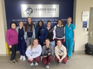 A group of ten people stand together infront of a Leader Dogs for the Blind sign smiling at the camera.