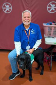 A man with white hair and a white beard is kneeling indoors. In front of him sits a black Labrador in a green Leader Dog ambassador vest. The man is smiling widely toward the camera while holding onto the dog. 